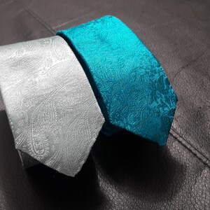 May include: Two patterned neckties, one silver and one teal blue, laid out on a dark gray textured surface.