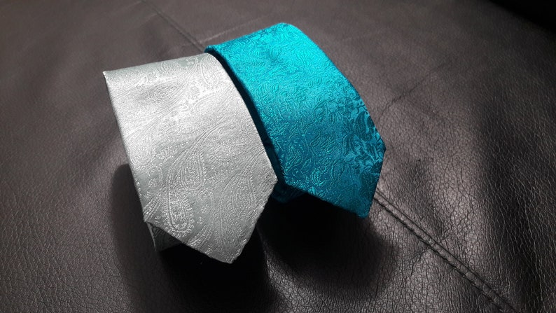 May include: Two patterned neckties, one silver and one teal blue, laid out on a dark gray textured surface.