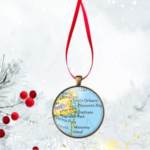 May include: Round Christmas ornament featuring a map of Cape Cod, with a diameter of 1.57 inches. The map is set in a bronze-colored frame and hangs from a red ribbon. The ornament includes place names like Harwich and Chatham.