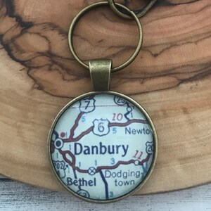 May include: A round, bronze-colored keychain pendant featuring a map of Danbury, with road numbers and the towns of Bethel and Dodgingtown. The pendant is attached to a bronze-colored key ring and clasp.