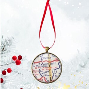 May include: Round Christmas ornament with a map design, featuring the text "Berlin" and "Gorham". The ornament has a bronze-colored metal frame and a red ribbon for hanging. The background is a soft, blurred white.
