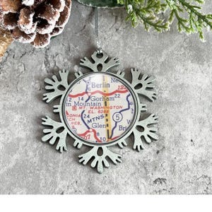 May include: A silver snowflake ornament with a map of Gorham, New Hampshire inside. The map shows the town's roads, landmarks, and the text "Gorham, New Hampshire".