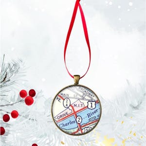 May include: Round Christmas ornament featuring a map design with a red ribbon for hanging. The map includes street names and numbers. The ornament has a bronze-colored metal frame and a red ribbon.