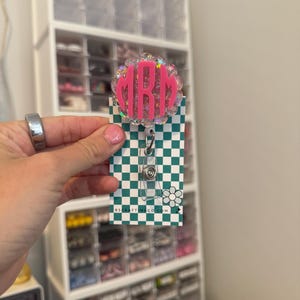 Peut inclure: Un porte-badge à monogramme rose et pailleté iridescent avec les initiales "MRM" sur fond blanc. La bobine est attachée à un porte-badge en plastique transparent avec un clip pivotant argenté. Le porte-badge est sur une carte à carreaux verts et blancs avec le texte "RSCRAFTINGCO.COM".