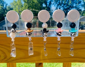 May include: Five black and white cow print retractable badge holders with different colored cowboy hats. The hats are turquoise, pink, black, brown, and white.
