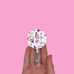May include: A round, pink and white striped retractable badge reel with a golf theme. The reel features illustrations of golf carts, golf bags, golf balls, and flags. The reel is held in a hand against a pink background.