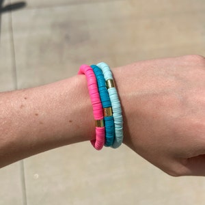 May include: Three stacked bracelets on a wrist. The top bracelet is light blue, the middle bracelet is teal, and the bottom bracelet is hot pink. Each bracelet has gold accents.