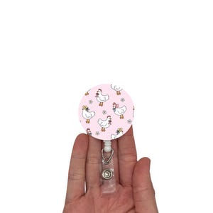 May include: A pink retractable badge reel featuring a pattern of cartoon chickens wearing hats and bows. The reel has a clear plastic strap and a metal clip.