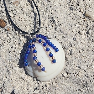May include: A handmade necklace featuring a white, rounded pendant adorned with strands of blue and clear beads. The beads are strung on silver-colored wire, and the necklace has a black cord. The pendant is resting on a sandy surface.