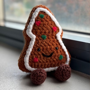 Puede incluir: Un hombre de jengibre tejido a mano, marrón, con ribete de glaseado blanco y chispas rojas y verdes. El hombre de jengibre tiene una cara sonriente y dos pies de bola tejidos marrones. Un juguete de peluche con temática festiva.