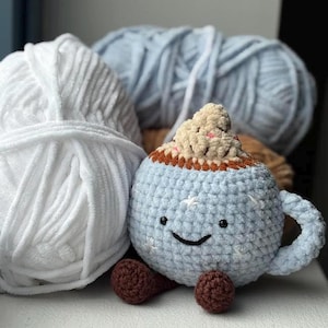Può includere: Una tazza da caffè blu all'uncinetto con un volto sorridente, piedini marroni e una guarnizione di panna montata. Sullo sfondo ci sono gomitoli di lana bianchi e blu. Peluche fatto a mano.