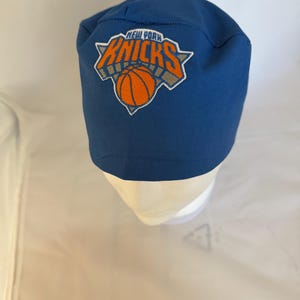 May include: A blue scrub cap featuring an embroidered New York Knicks basketball team logo on the front. The logo displays the text New York Knicks in orange and white lettering above an orange basketball graphic with a grey triangular background.