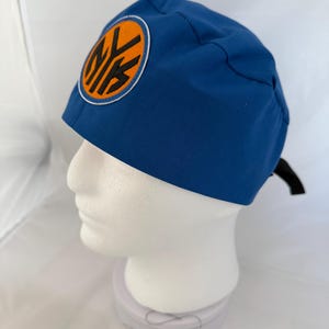 May include: A royal blue scrub cap featuring a circular patch on the side. The patch has an orange background with a white border and the black letters NY in a stylized font. The cap is secured with a black tie at the back of the head.