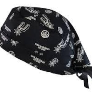 May include: A black scrub cap featuring a repeating white pattern of the San Antonio Spurs basketball team logo and the text San Antonio Spurs. The cap has a folded front band and black fabric ties at the back for an adjustable fit.