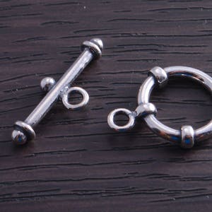 May include: Two silver metal clasps for jewelry making. One clasp is a toggle clasp with a round bar and a loop. The other clasp is a round ring with a small loop on one side.