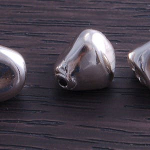 May include: Three silver-colored metal beads, each with a unique shape, are arranged in a row on a dark brown wooden surface.