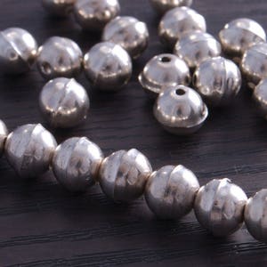 May include: A close-up of a string of silver beads with a textured surface. The beads are arranged in a line and are all the same size. The beads are on a dark brown surface.