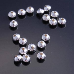 May include: A collection of small, shiny silver-colored metal bead caps with a rounded, conical shape and a small hole at the top. The beads are scattered on a dark surface, reflecting light.