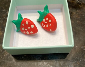 May include: A pair of red and green strawberry-shaped earrings in a light blue box. The earrings are made of polymer clay and have white dots on them.