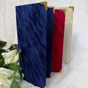 May include: Three tall, rectangular menu covers standing upright. The first is dark blue, the second is deep red, and the third is cream. Each cover features a textured fabric finish and decorative gold-toned metal corner protectors.