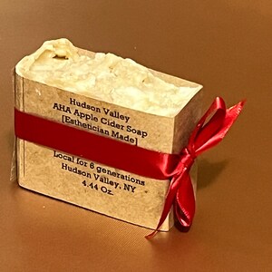 May include: A brown paper wrapped bar of soap with a red ribbon tied around it. The soap is labeled "Hudson Valley AHA Apple Cider Soap [Esthetician Made]" and "Local for 6 generations Hudson Valley, NY 4.44 Oz."