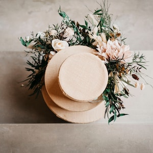 May include: A decorative arrangement featuring three stacked, round, beige burlap-covered platforms. The platforms are adorned with a variety of flowers, including white roses and a large, light pink dahlia, and greenery. The arrangement is set against a neutral background.