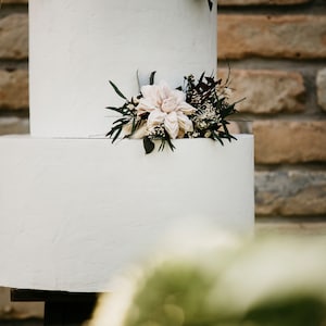 May include: A two-tiered white cake with a textured finish, adorned with a floral arrangement of light pink and dark green foliage. The cake is set against a rustic brick wall, creating a contrast of textures and colors.