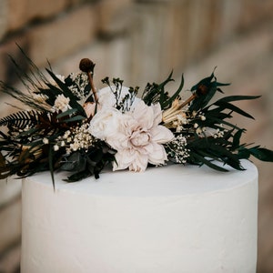 May include: A white, tiered cake topped with a floral arrangement. The arrangement features a large, light pink flower, dark green leaves, and small white flowers. The cake is set against a blurred brick background.