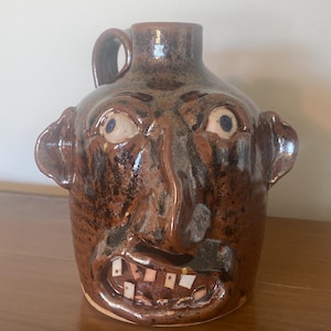 May include: A brown ceramic jug with a face sculpted on the front. The jug has a handle and a spout. The face has bulging eyes, a large nose, ears, and a toothy grin. The jug is glazed with a glossy finish.