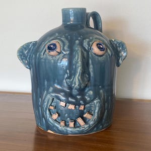 May include: A blue ceramic face jug with a glossy finish, featuring a sculpted human-like face with protruding ears, a prominent nose, and a wide, open mouth displaying individual light-colored teeth. The vessel has a single handle on the back.