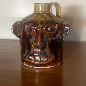 May include: A brown ceramic jug with a face design. The jug features a handle, a spout, and a detailed face with eyes, a nose, a mouth, and ears. The colors include shades of brown and tan, with a glossy finish. The jug is a decorative item.