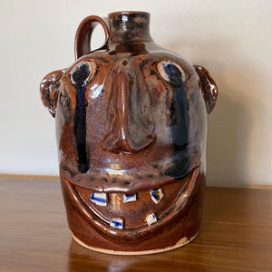 May include: A brown glazed ceramic face jug with a handle, featuring a wide smiling mouth with individual white and blue teeth, prominent ears, and dark blue glaze running from the eyes down the cheeks, creating a distinct character design.