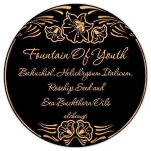 May include: A circular black label with a thin gold border and gold floral accents at the top and bottom. The text reads: Fountain Of Youth, Bakuchiol, Helichrysum Italicum, Rosehip Seed and Sea Buckthorn Oils, and the word alchemy6 in gold script.