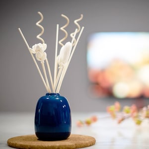 May include: A blue ceramic diffuser bottle with white wooden sticks and white flower-shaped diffusers. The diffuser is sitting on a cork coaster.