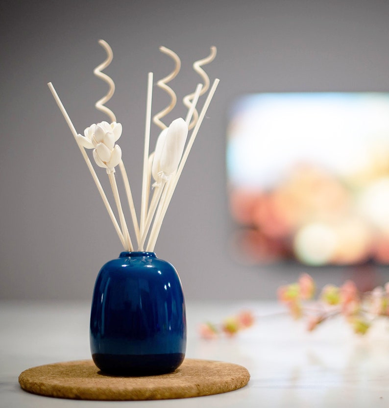 May include: A blue ceramic diffuser bottle with white wooden sticks and white flower-shaped diffusers. The diffuser is sitting on a cork coaster.