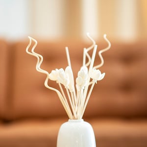 May include: White ceramic vase with a bouquet of white wooden flowers and brown sticks. The vase is sitting on a light brown wooden table.