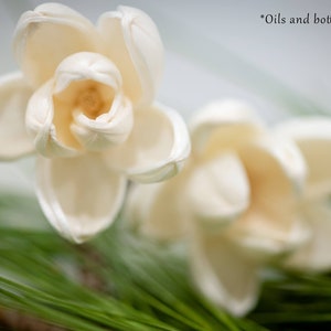 May include: Two white, paper-like flowers with a delicate, folded texture are nestled in a bed of green pine needles. The flowers are arranged in a natural, organic style, creating a simple and elegant display.