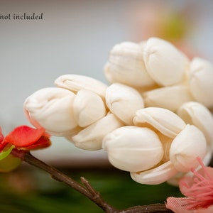 May include: A close-up of a bouquet of white artificial flowers with a stem and a small pink flower in the background.