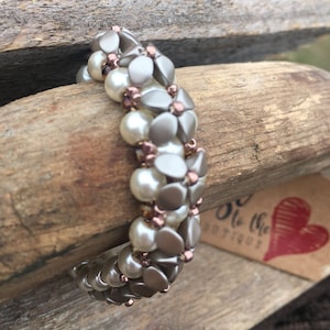 Puede incluir: Un brazalete de cuentas beige y marrón con un diseño floral. El brazalete está hecho con cuentas pequeñas y redondas y cuentas más grandes y planas que se asemejan a pétalos de flores.