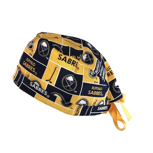 May include: A yellow and blue surgical scrub hat with a Buffalo Sabres NHL team logo pattern. The hat has a tie on the side.