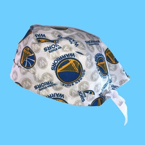 May include: White fabric scrub hat with a repeating pattern of the Golden State Warriors logo. The logo features a basketball with a blue and yellow design and the words "Golden State Warriors" in blue text.
