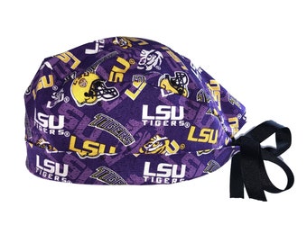 May include: A purple scrub cap featuring a repeating pattern of the LSU Tigers logo, yellow LSU text, and yellow football helmets. The cap has a wide band and black fabric ties at the back to secure the fit, showcasing a collegiate sports theme.