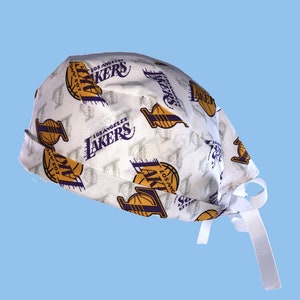 May include: White fabric scrub hat with a repeating pattern of the Los Angeles Lakers logo in purple and gold. The hat has white ties.