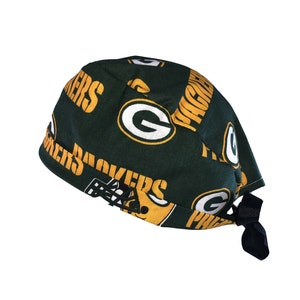 May include: Green and gold fabric scrub hat with the Green Bay Packers logo and the words "Packers" repeated on the fabric. The hat has a black tie.
