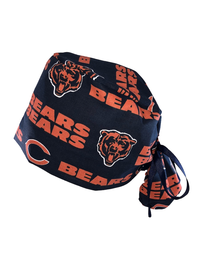 May include: A navy blue surgical scrub hat with a tie. The hat is covered in a repeating pattern of orange bears and the word "Bears".