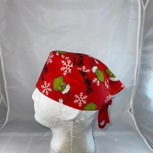May include: Red and green surgical scrub hat with a Christmas theme. The hat features a repeating pattern of snowflakes and the Grinch character. The text "Grinchmas" is printed on the hat.