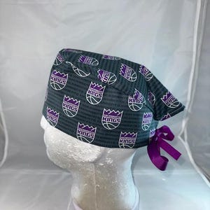 May include: A purple and gray scrub hat with a Sacramento Kings basketball team logo pattern. The hat has a purple tie on the back.