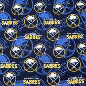 May include: A navy blue fabric featuring a repeating pattern of the Buffalo Sabres logo and wordmark. The logo includes a white bison head within a gold circle, crossed hockey sticks, and the text "BUFFALO SABRES" in gold.