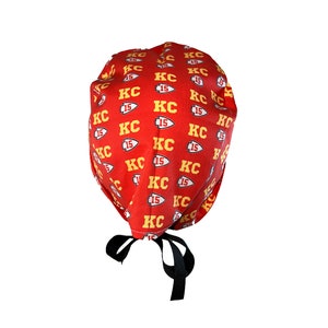 May include: Red surgical scrub hat with a repeating pattern of yellow "KC 15" logos. The hat has black ties.