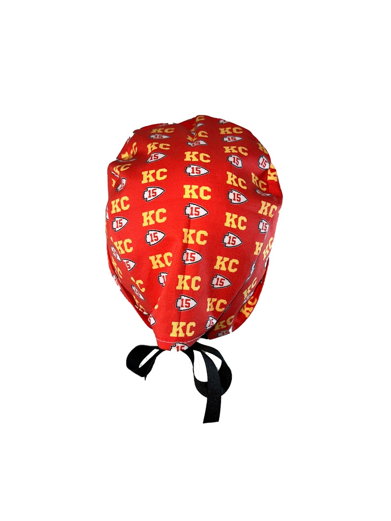 May include: Red surgical scrub hat with a repeating pattern of yellow "KC 15" logos. The hat has black ties.